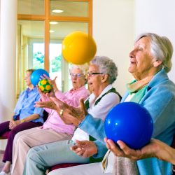 Group of happy senior ladies doing coordination exercises in a seniors gym sitting in chairs throwing and catching brightly colored balls.