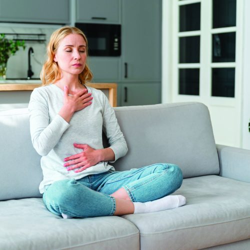 Mindful young woman with hands on chest and stomach breathing out with closed eyes, calming down at home in stressful situation. Managing stress, reaching inner balance.